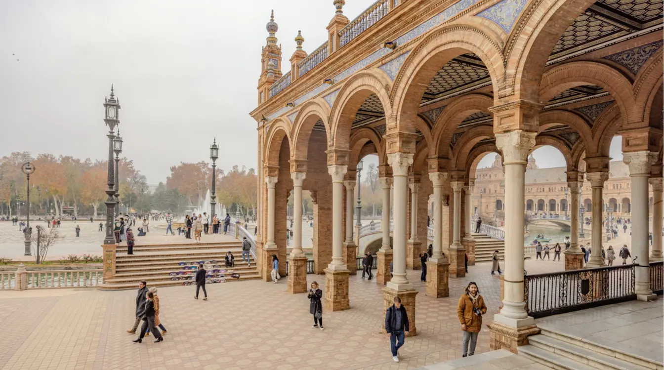 Vista da Plaza de España, em Sevilha, com moradores e viajantes caminhando pela calçada e as árvores com folhas em queda no fundo.