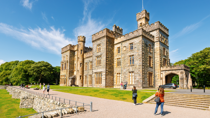 The sand-coloured Lews Castle, with its Gothic turrets, archways and stairwells, against a blue sky and lush green gardens on Stornoway in Scotland. 