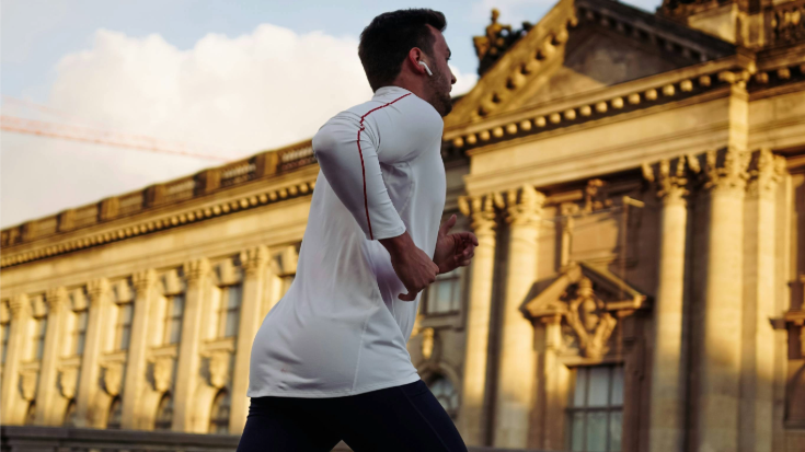 A colour photograph of a young man of asian descent with olive skin and black hair and beard dressed in a white running shirt and black running shorts. He is running past Berlin's famous Reichstag building. To illustrate a blog post entitled 'Berlin Half Marathon 2026​: Can You Still Register?'