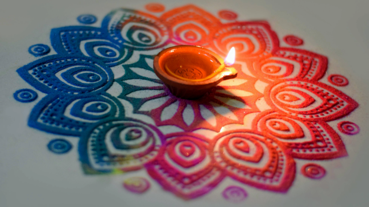A traditional Indian rangoli pattern in orange, blue, pink and green with a lit candle in the centre. 