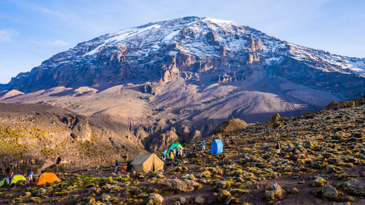 A view of the snow capped peak of Mount Kilimanjaro in Tanzania from the town of Moshi. It shows a group of people walking towards the mountain as they make their way through a boulder field, to illustrate a blog post entitled 'The most beautiful half-marathons in the world'.