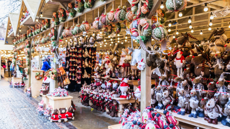 An image of Alpine style wooden huts at an outdoor Christmas market in the UK. Christmas decorations such as baubles in greens, reds and whites hang from strings and the huts stretch far along a cobbled square. To illustrate a blog post entitled 'Things to do in London in December'. 