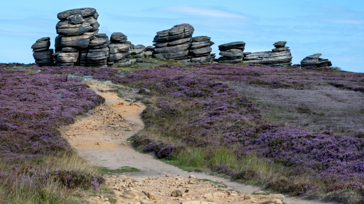 A dramatic rock formation in the Peak District, showing layered textured rock towers sitting on the edge of rolling hills.