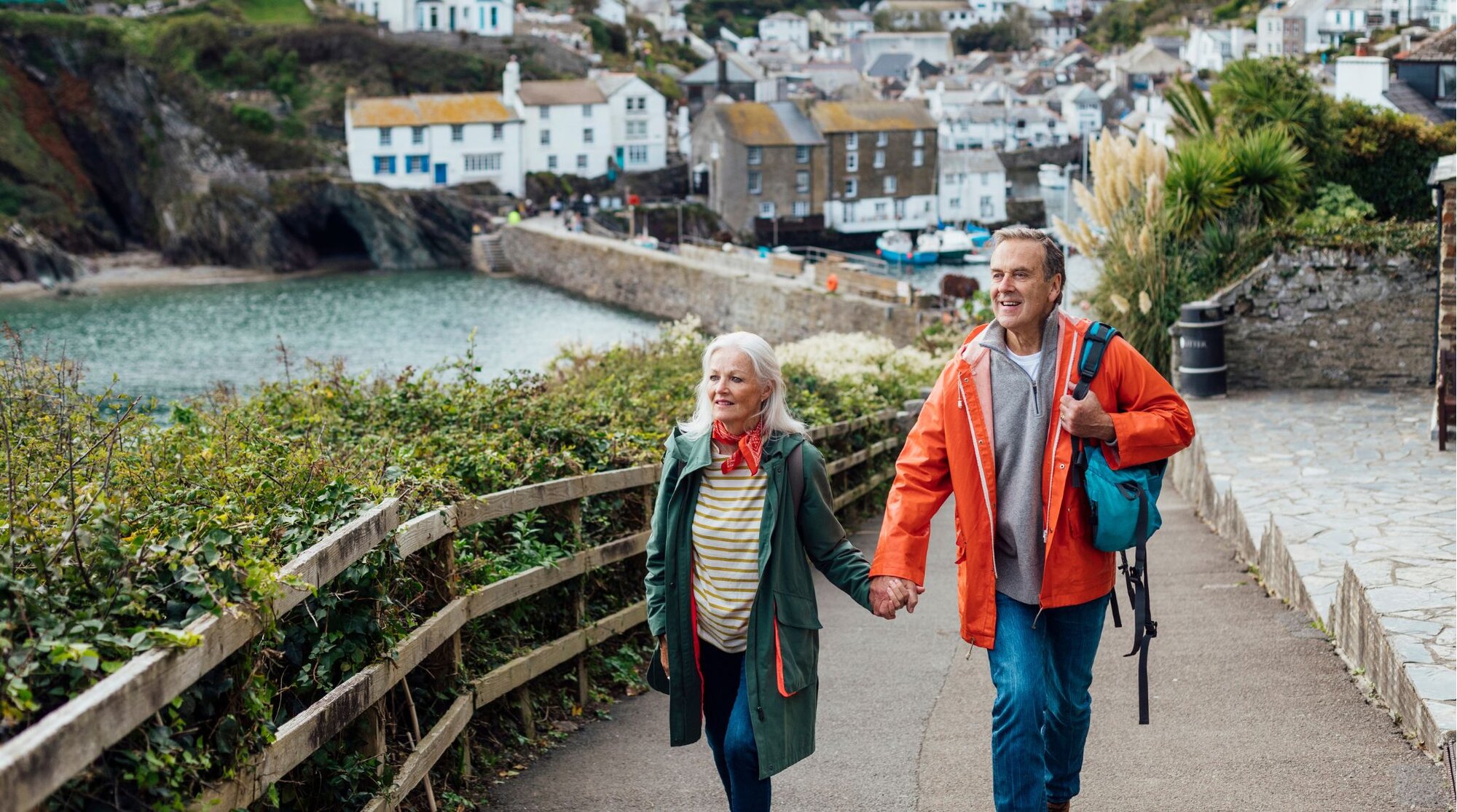 Casal de meia-idade caminhando de mãos dadas em vilarejo costeiro europeu, com mochilas e casacos coloridos.