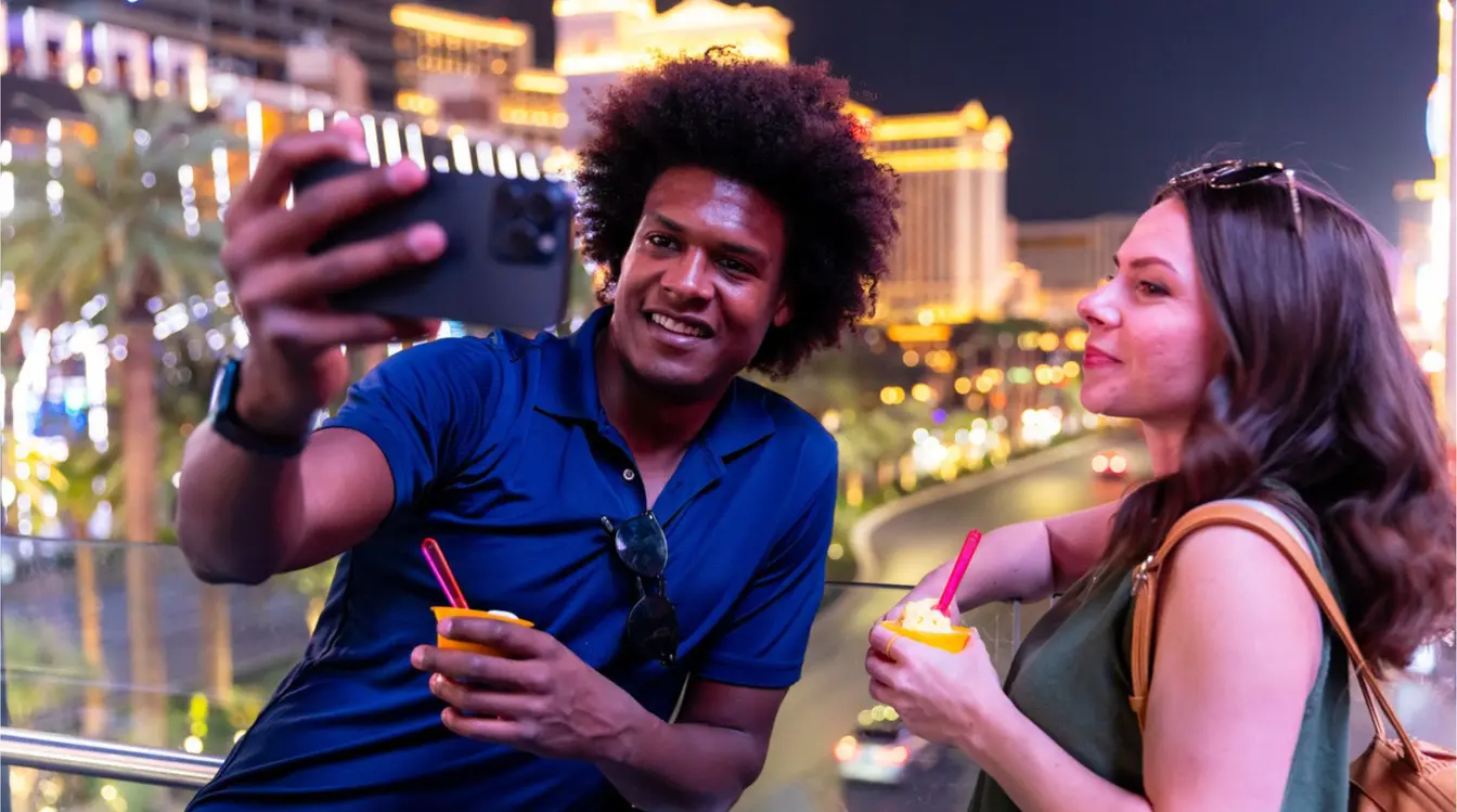Pareja tomándose una selfie de noche en un puente peatonal, con el Strip iluminado al fondo y bebidas en la mano.
