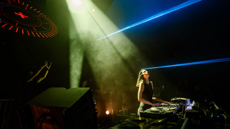 A photograph of a DJ. She is in the foreground and tilting her head back, immersed in her music wearing headphones and adjusting her deck. The background is completely dark, except for one spotlight, which is illuminating the DJ. To illustrate a blog post entitled 'The Ultimate Sziget Festival Guide.'