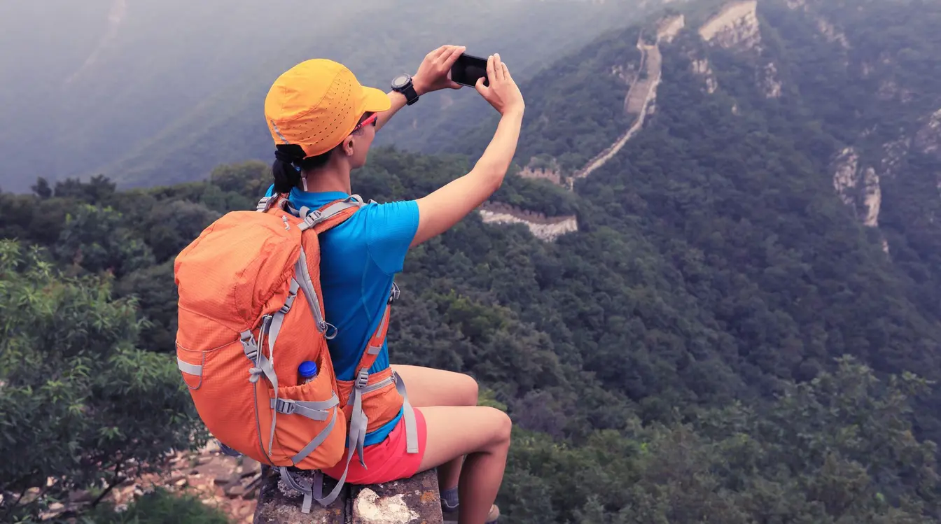 A woman sat on a ledge, using a smartphone to take a picture of the Great Wall of China and the surrounding forested hills.