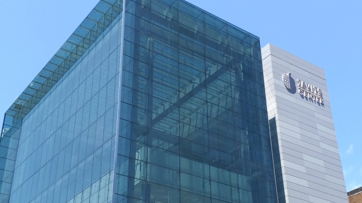 A colour photograph of the Javits convention Center in West Side Manhattan, New York City. The building is made a of glass and steel, is grey and transparent and states 'JAVITS CENTER' on thie side via. a large sign. In the background, the sky is bright blue and clear of clouds and light is reflecting off the windows of the convention center. To illustrate a blog post entitled 'New York Comic Con 2025: A Complete Travel Guide for Attendees .'