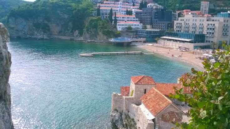 A photograph of a beach in Budva, Montenegro, from the top of a cliff, showing a quiet atmosphere with a few holidaymakers walking on a sandy yellow beach. In the background are more clifs and an apartment complex, to illustrate a blog post by the eSIM company Airalo entitled 'Where is hot in October in Europe?'