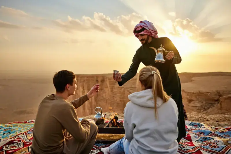 Hombre sirviendo té a una pareja sentada al aire libre en un paisaje desértico al atardecer.