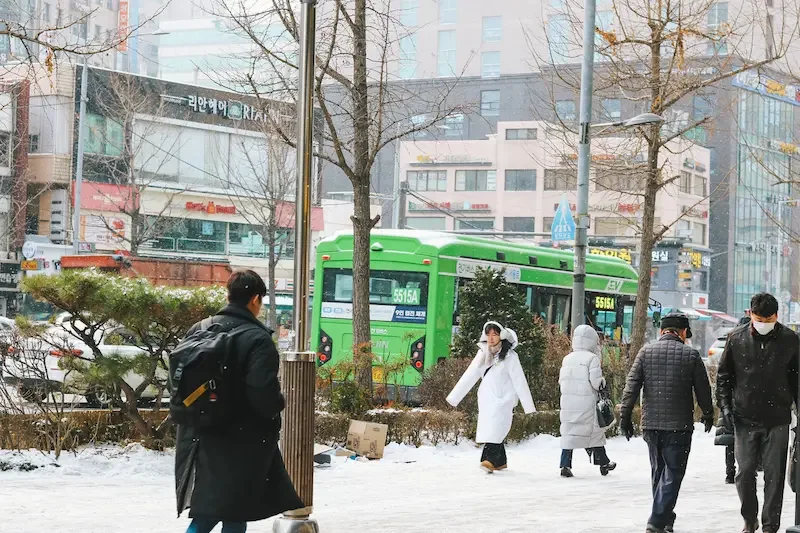 雪の積もる冬のソウルの街並みと、厚手のコートを着て歩く人々や緑色の路線バス