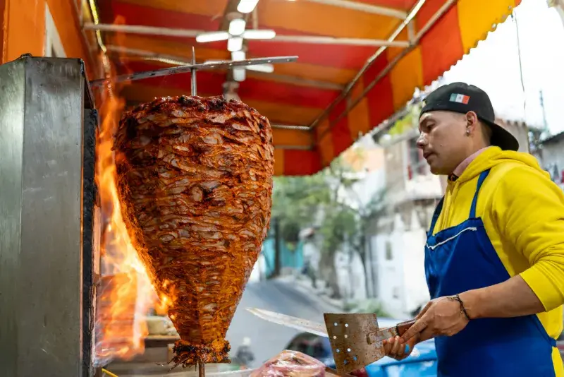 Taquero preparando carne al pastor en un trompo en un puesto callejero.
