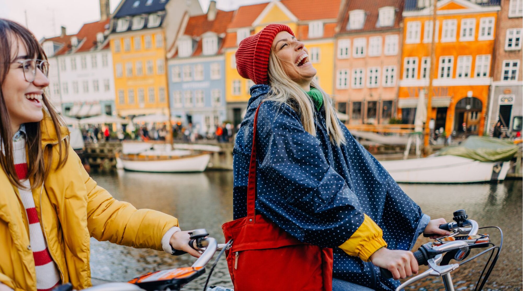 Duas amigas andando de bicicleta e sorrindo às margens do canal de Nyhavn, em Copenhague, Dinamarca.