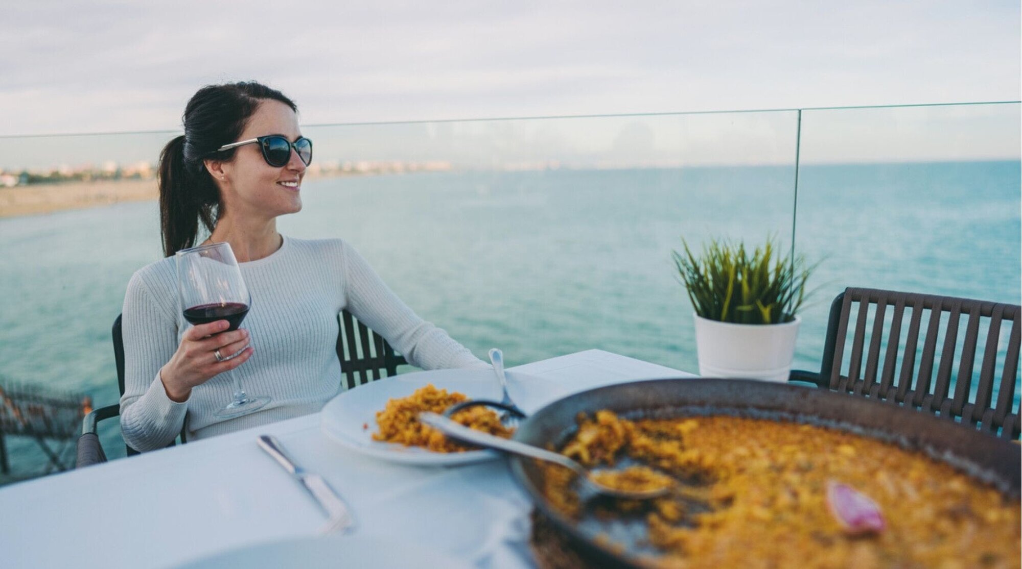 Mulher em um restaurante comendo a tradicional paella espanhola, acompanhada de uma taça de vinho tinto.
