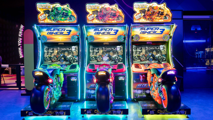 An image of three game arcade units at the Esports World Cup 2025, each has a large motorbike in front of a gaming screen and they are all lit in bright colours.