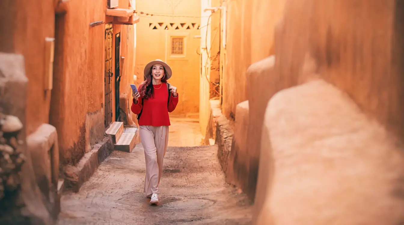 Junge Touristin mit Rucksack und Hut nutzt mobile Daten im Ausland in einer Gasse von Ait Benhaddou, Marokko.