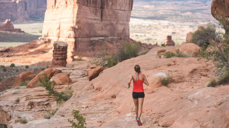 A female in her 20s jogs through the Utah desert as part of her training for an upcoming race. There are large rock formations rising in the desert in front of her. The shot is from behind.