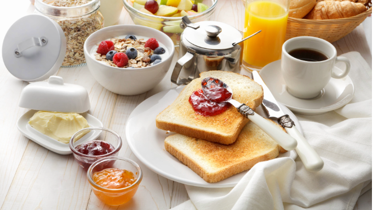 An image of a continental breakfast, featuring muesli in a glass jar, marmalade, jam, white toast, butter resting on a white butter dish and a glass of orange juice.