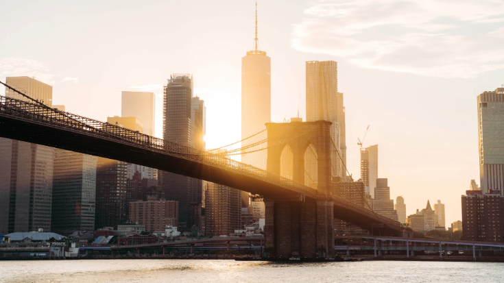 A sepia coloured photograph with flare of New York City at sunset. It shows a view over Manhattan, with the Brooklyn bridge in view and the World Trade center in the background 