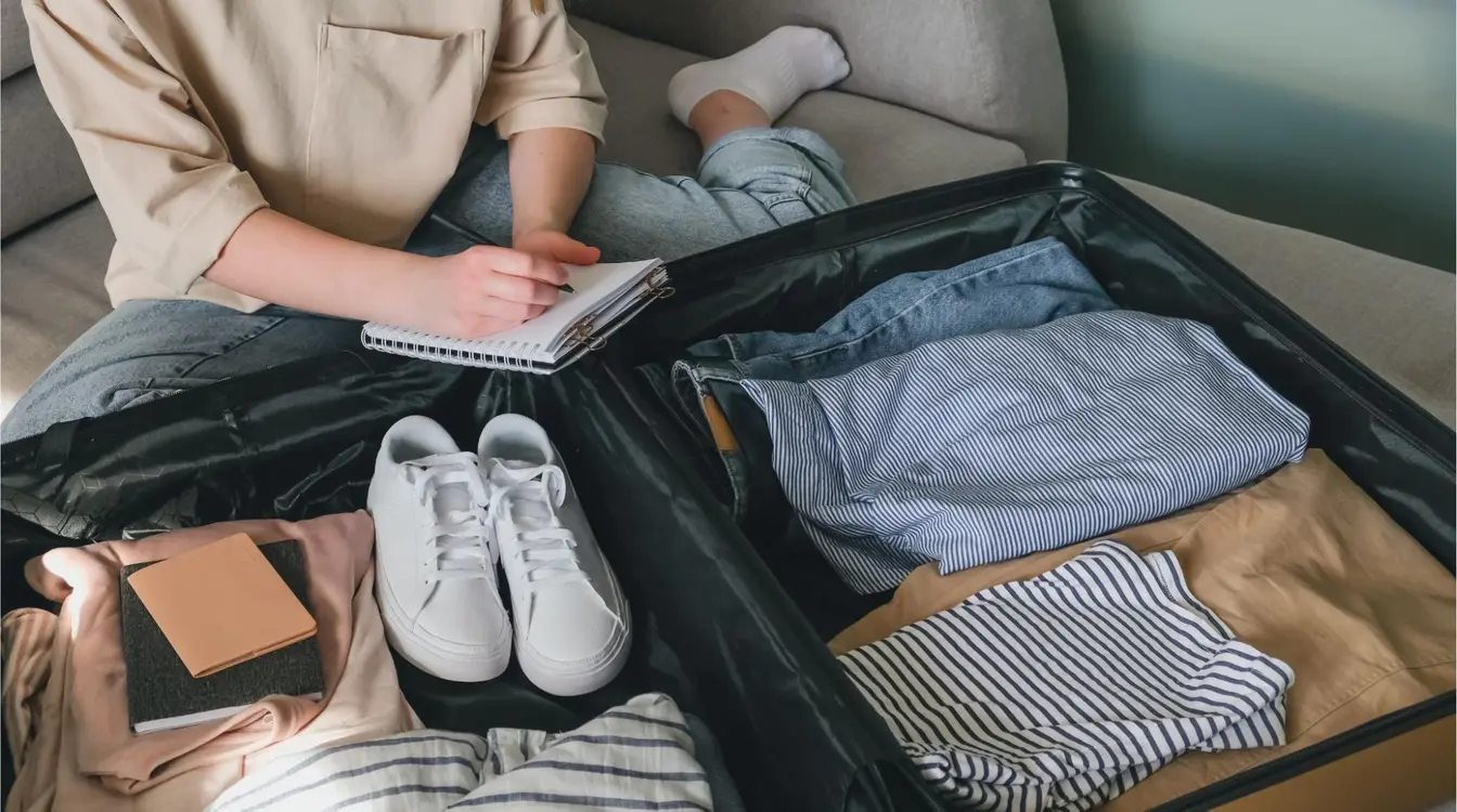 Uma pessoa sentada no sofá ao lado de uma mala de viagem aberta com roupas, tênis brancos e uma carteira, fazendo um check list para viagem internacional.