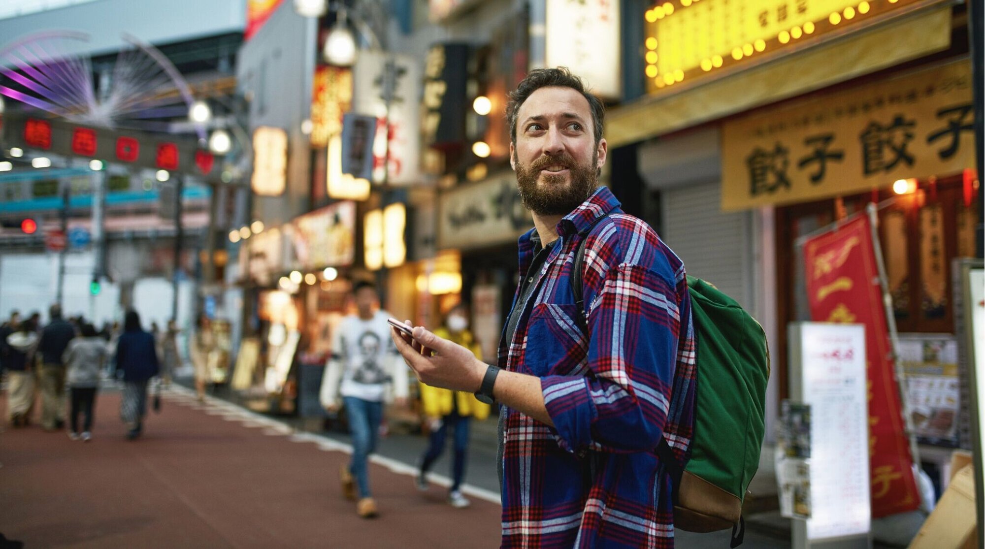 Viajante com mochila usando o celular em rua iluminada de Tóquio, conectado com chip internacional Japão.