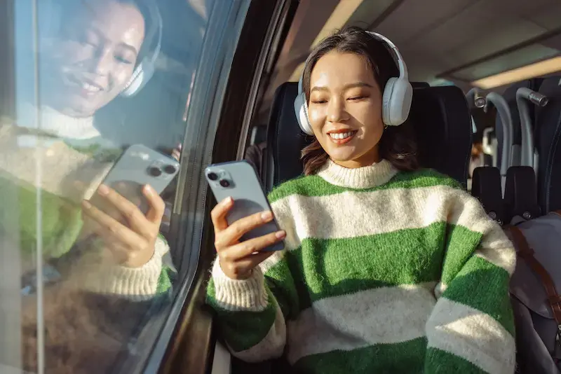 Una ragazza in treno al finestrino sorride al suo smartphone indossando le cuffie.