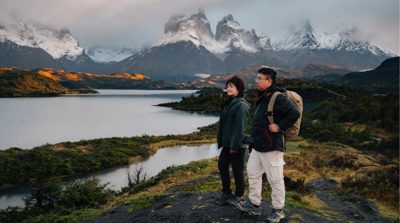 Casal de turistas com roupas de frio e mochilas em pé perto de um lago, admirando uma paisagem imponente com montanhas de picos nevados ao fundo, característica da região da Patagônia.