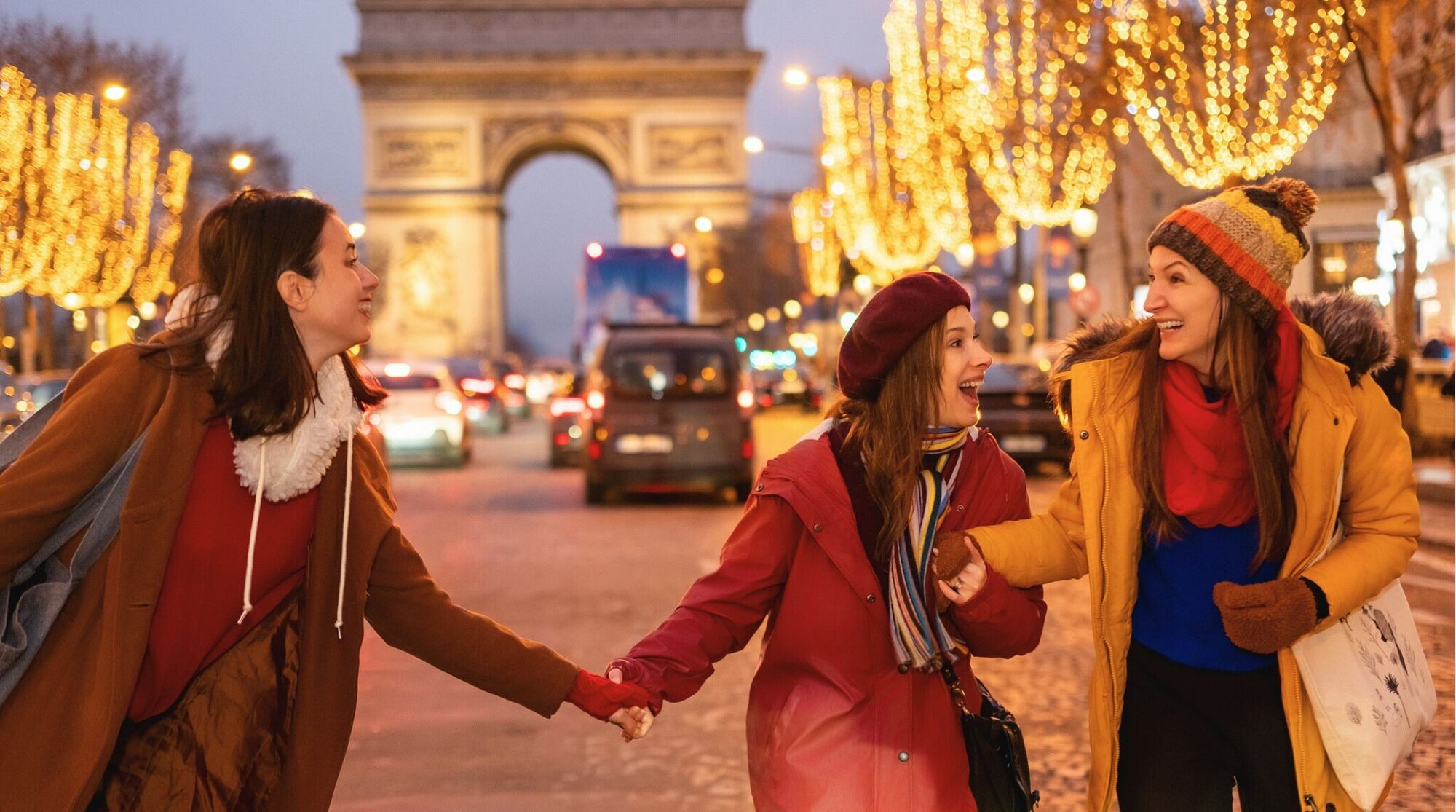 Três mulheres com roupas de inverno riem e dão as mãos enquanto caminham por uma rua com decoração natalina, com o Arco do Triunfo ao fundo.