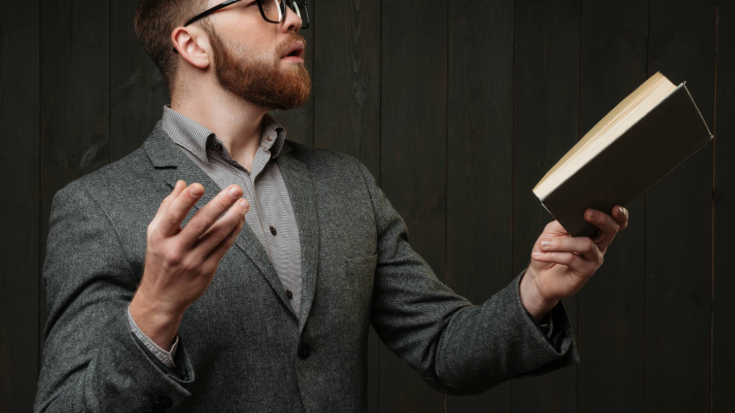 A colour image of a man against a dark background. He is dressed in a light grey shirt and a dark grey jacket and has a ginger beard and closely cut ginger hair. He has thick plastic framed black glasses. He is looking over to his left and appears to be addressing a room of people and reciting a poem from a book he is holding open in his left hand. To illustrate a blog post entitled 'What is Burns Night and How to Celebrate It'.