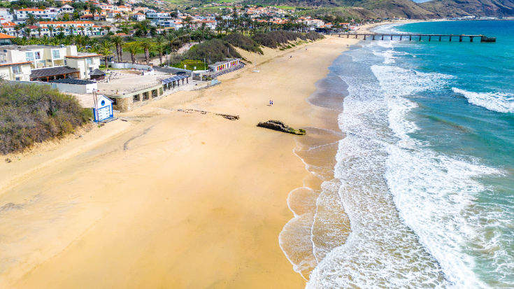 An aerial view of yellow sandy Porto Santo Beach, a popular beach on the Portuguese Island in the Atlantic Ocean. In the background are buildings of Vila Baleira, to illustrate a blog post entitled 'Where is hot in October in Europe?'