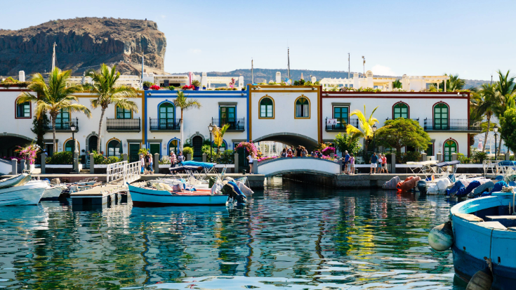 A photograph of Puerto de Morgan on Gran Canaria Island, Spain. In the foreground is a body of blue water and int he background is a row of whitewashed houses, with blue and brown trims painted around their windows and doors. There are people dressed in summer clothes walking in front of the houses and enjoying the views across the water. There are also green leaved palm trees at the front of some of the houses and the sky is a very clear blue. To illustrate a blog post entitled 'Where Is Hot in November in Europe?'