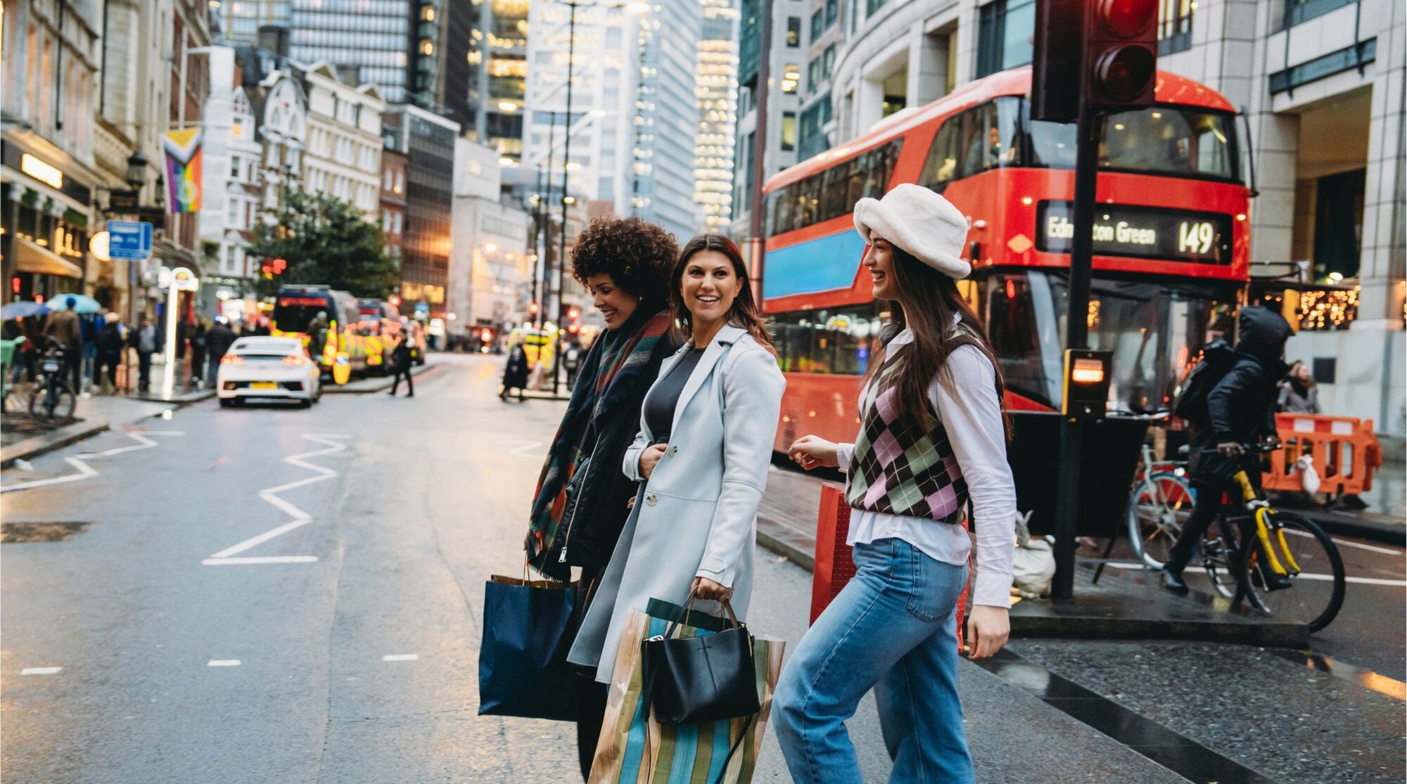Trio de amigas viajantes em Londres, caminhando pelas ruas com tradicional ônibus vermelho no fundo.