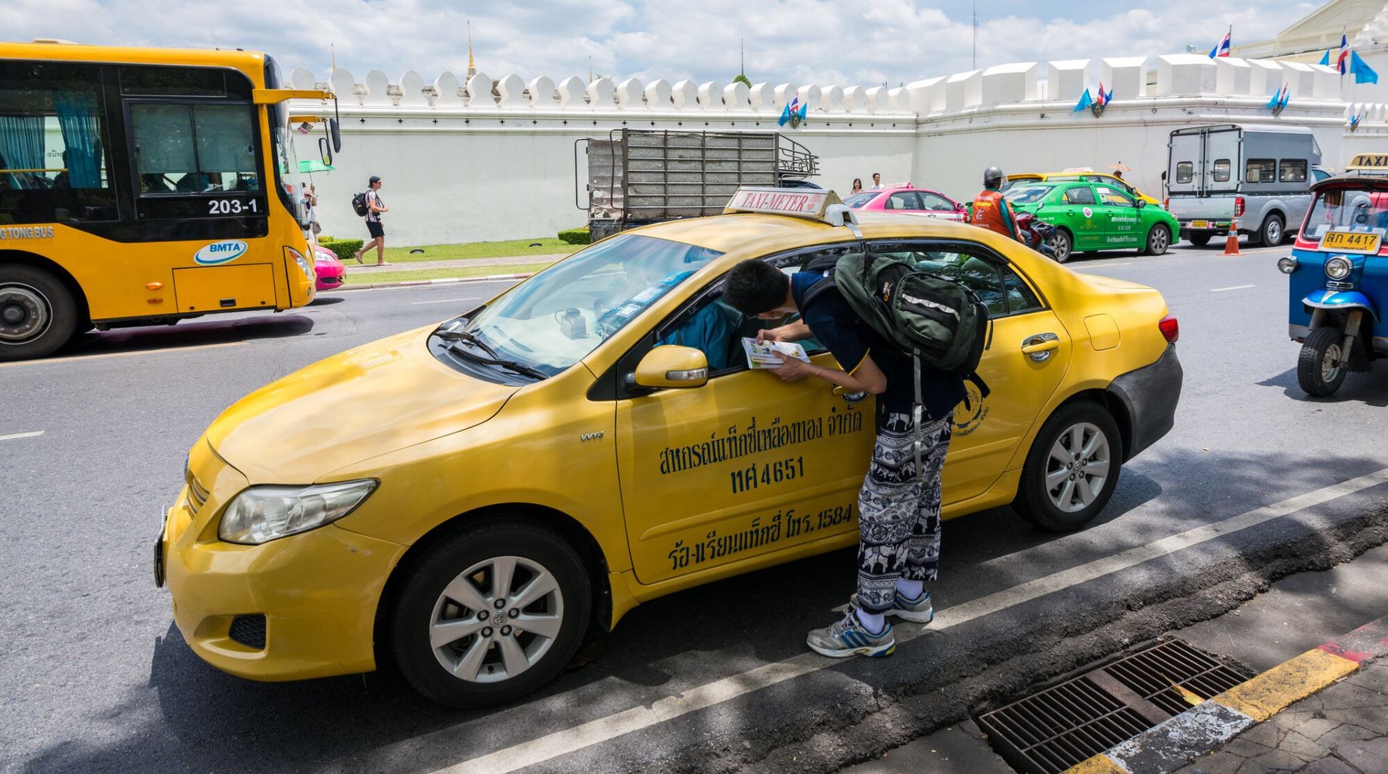 A tourist leaning toward the window of a yellow metered taxi to talk to the driver on a street in Bangkok.