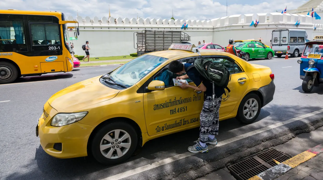 A tourist leaning toward the window of a yellow metered taxi to talk to the driver on a street in Bangkok.
