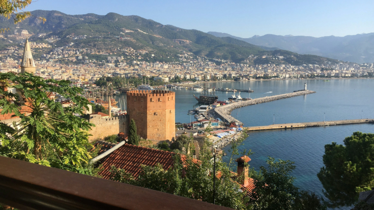 A photograph showing a view of a red-walled castle Red Tower, with Alanya harbour and the Taurus Mountains in the background - one of the top Alanya attractions for couples and families, to illustrate a blog post entitled things to do in Alanya.
