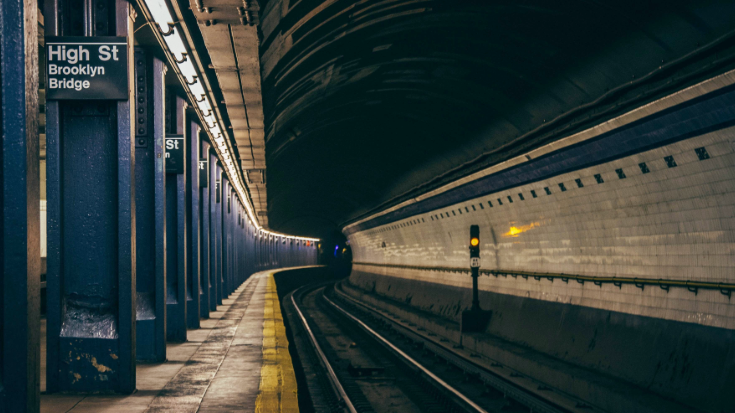A photograph of an underground station stop on the New York subway. The image shows an empty train line and platform, with a sign stating 'High St Brooklyn Bridge'. It shows The High Street stop in Brooklyn, served by the A and C trains and is a convenient subway station for accessing the Brooklyn Bridge.