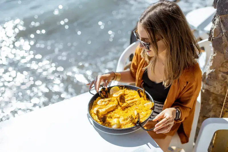 Mujer comiendo paella frente al mar en un restaurante al aire libre en España.