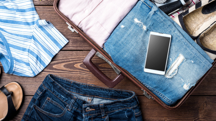 An image of clothes, including blue denim jeans, a pink short and a striped blue and white shirt, in and around an open suitcase on a wooden floor, with an iPhone thrown on top of the half-packed suitcase of clothes, to illustrate a blog post entitled 'Is My iPhone eSIM-Compatible?'