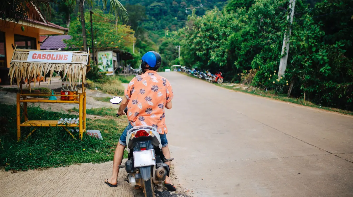 Rear view of a person on a motorbike stopped at a roadside gasoline stand in a rural setting.