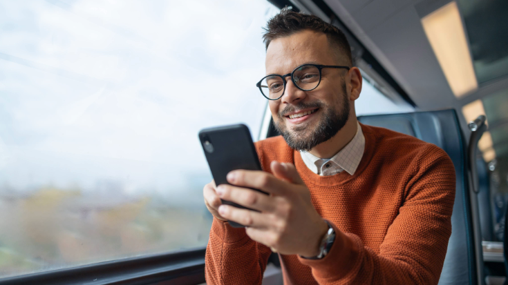 A man in an orange jumper travelling on a train while looking at his phone screen and smiling. To illustrate a blog post entitled '11 WhatsApp Text Tricks to Make Your Messages Pop.'