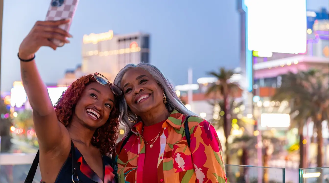 Dos mujeres tomándose una selfie en un mirador del Strip de Las Vegas por la noche, con luces de hoteles y tráfico iluminado al fondo.