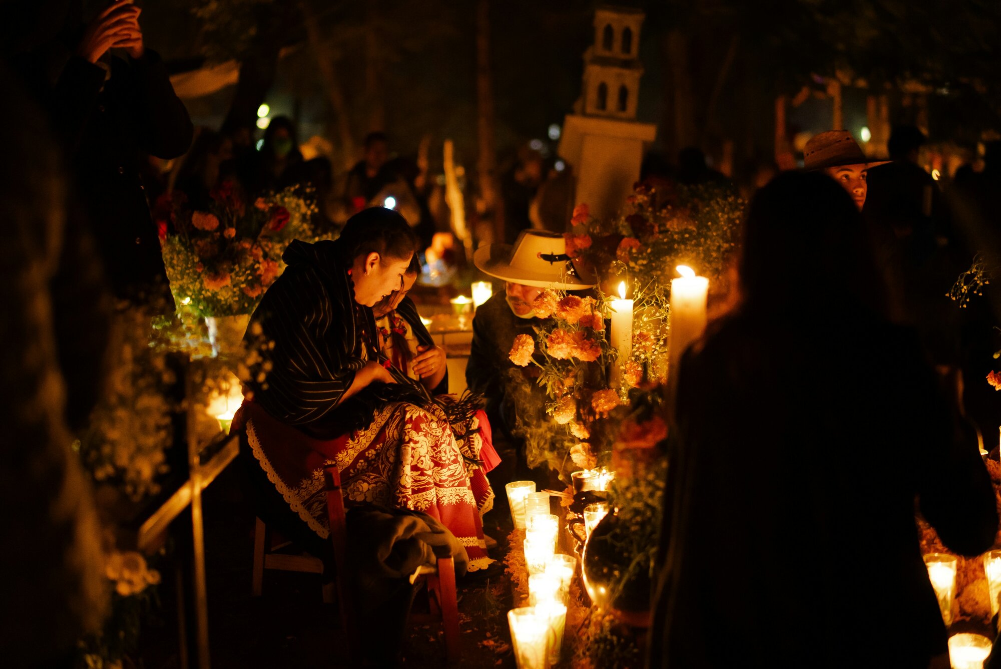 Imagen de un cementerio durante la celebración del Día de Muertos, donde varias personas con indumentaria tradicional se reúnen junto a tumbas iluminadas con velas