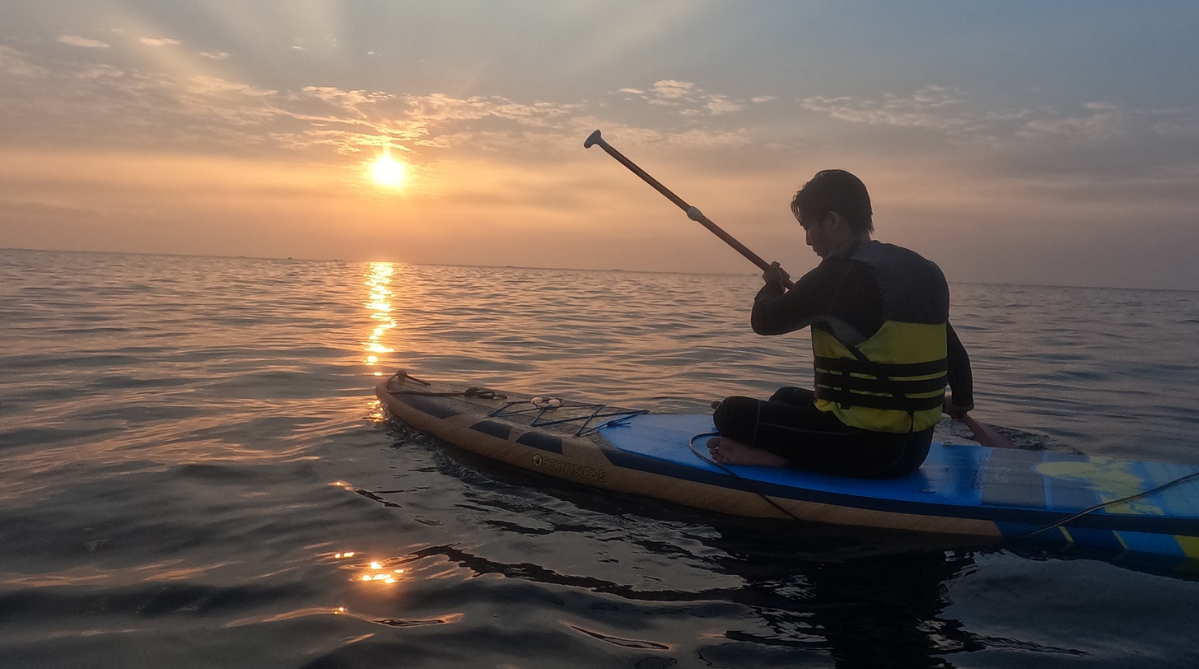 Man in wetsuit sitting on a standup paddleboard rowing into the sunrise horizon
