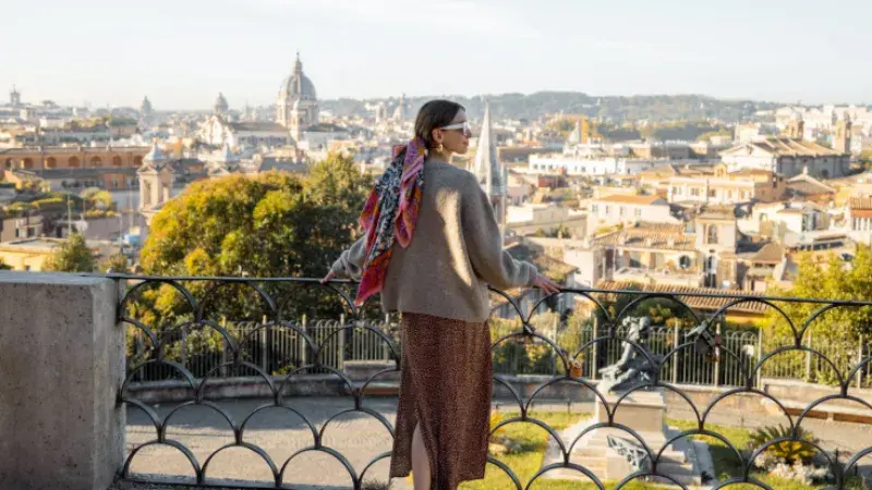 A brown haired woman wearing a colourful headscarf and sunglasses, looking out over a cityscape of Rome, Italy.
