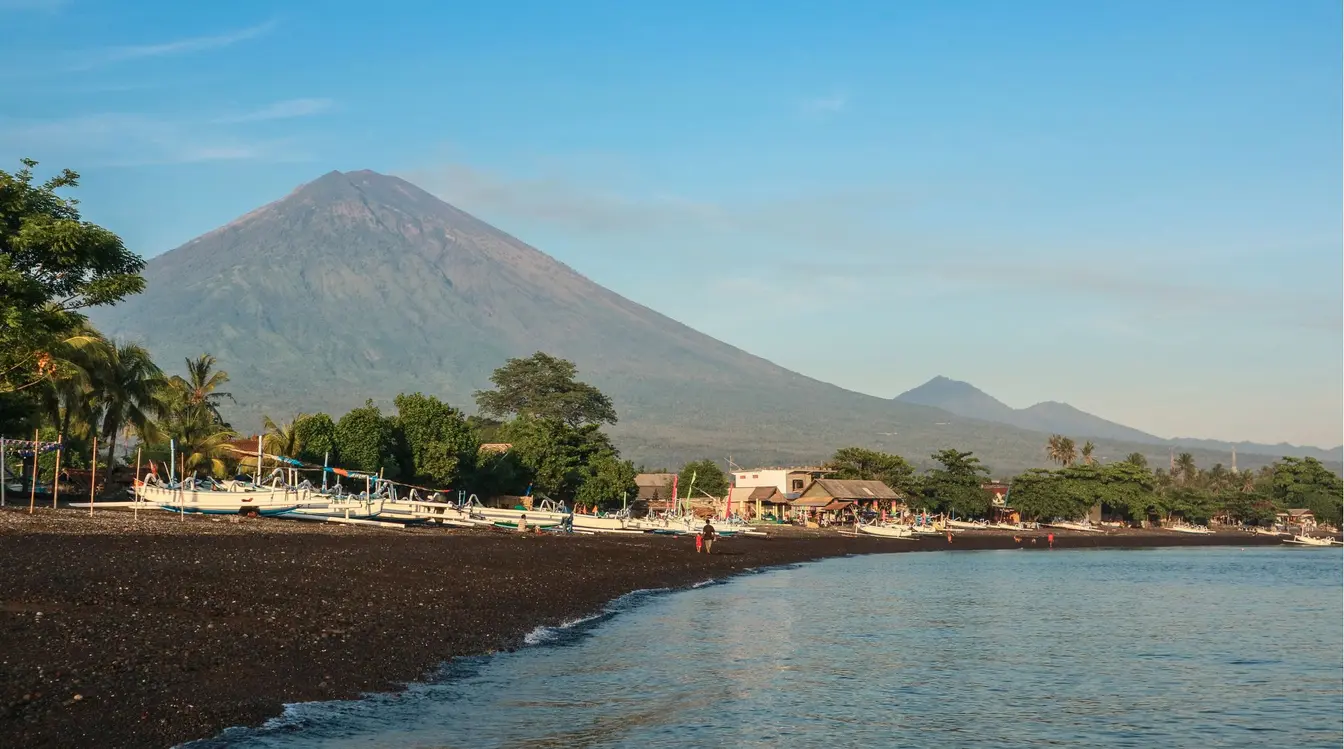 View of Mount Agung towering above a black sand beach in Amed, with boats lined up along the shore.