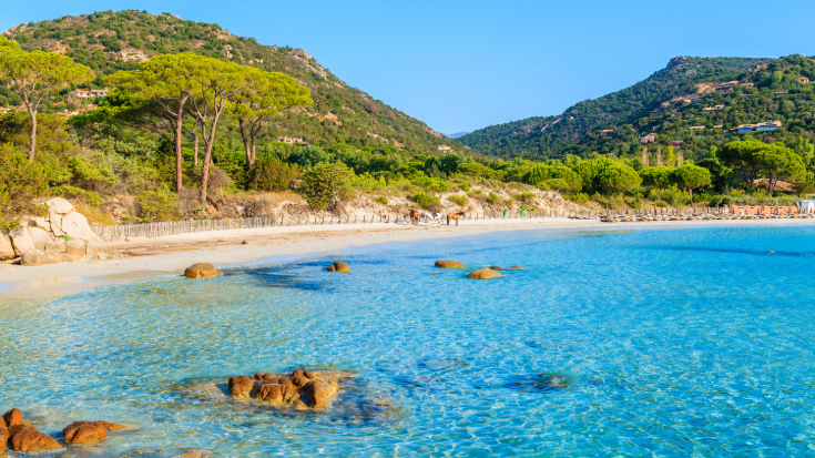 A photograph showing Palombaggia beach in Corsica, France. The beach curves up the coastline and has white sands, while in the background are some bright green hills. In the foreground is a sea shore with azure waters and sandy rocks. To illustrate a blog post entitled 'Where is Hot in December in Europe?'