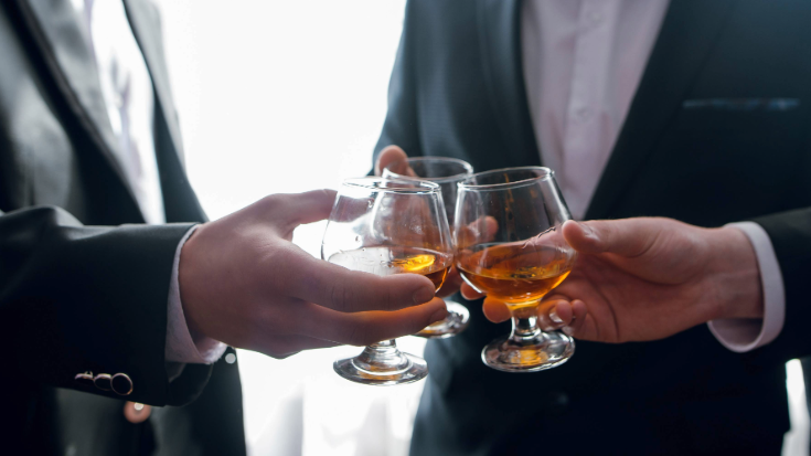 a colour image of three men's hands holding traditional tulip-shaped Scottish whiskey glasses, each filled about an eighth of the way with golden Scottish whiskey. To illustrate a blog post entitled 'What is Burns Night and How to Celebrate It'.