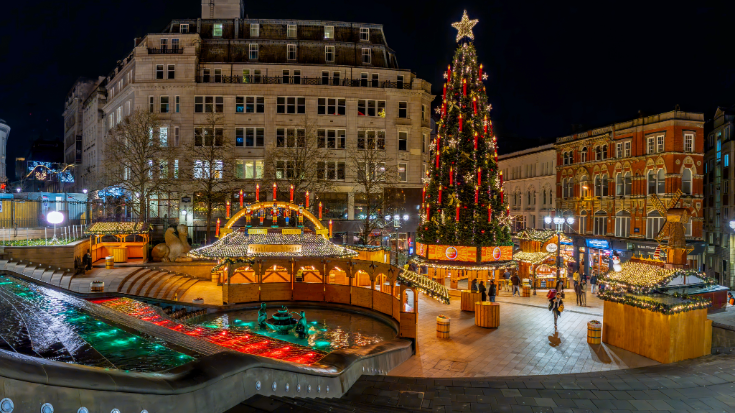A coloured image showing the Frankfurt Christmas Market in Birmingham, with wooden huts and Christmas decorations. In the landscape is a large outdoor Christmas tree, decorated with large red candles and glowing warm lights. In the image is also a fountain, surrounded by sandy stone steps, which are lit by blue spotlights. To illustrate a blog post entitled 'Extreme Day Trips - UK Christmas Markets Edition'.
