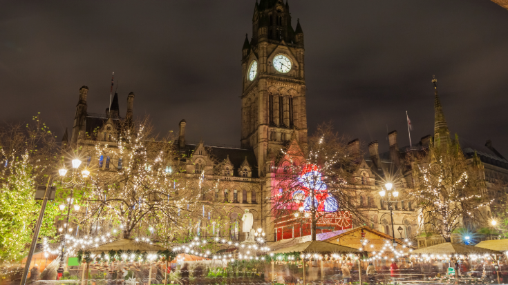 A colour photograph of a dark night sky in the background and Manchester's town hall in the foreground, complete with a large tall tower and an illuminated clock face. Under the town hall in the main square are lots of wooden Christmas market huts and lights illuminating the scene. To illustrate a blog post entitled 'Extreme Day Trips - UK Christmas Markets Edition'.