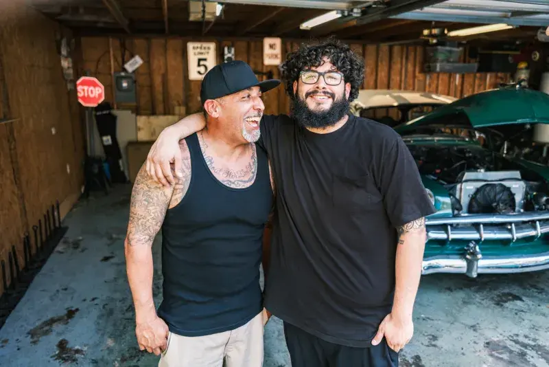 Dos hombres abrazados posando dentro de un garaje, con un coche clásico al fondo.